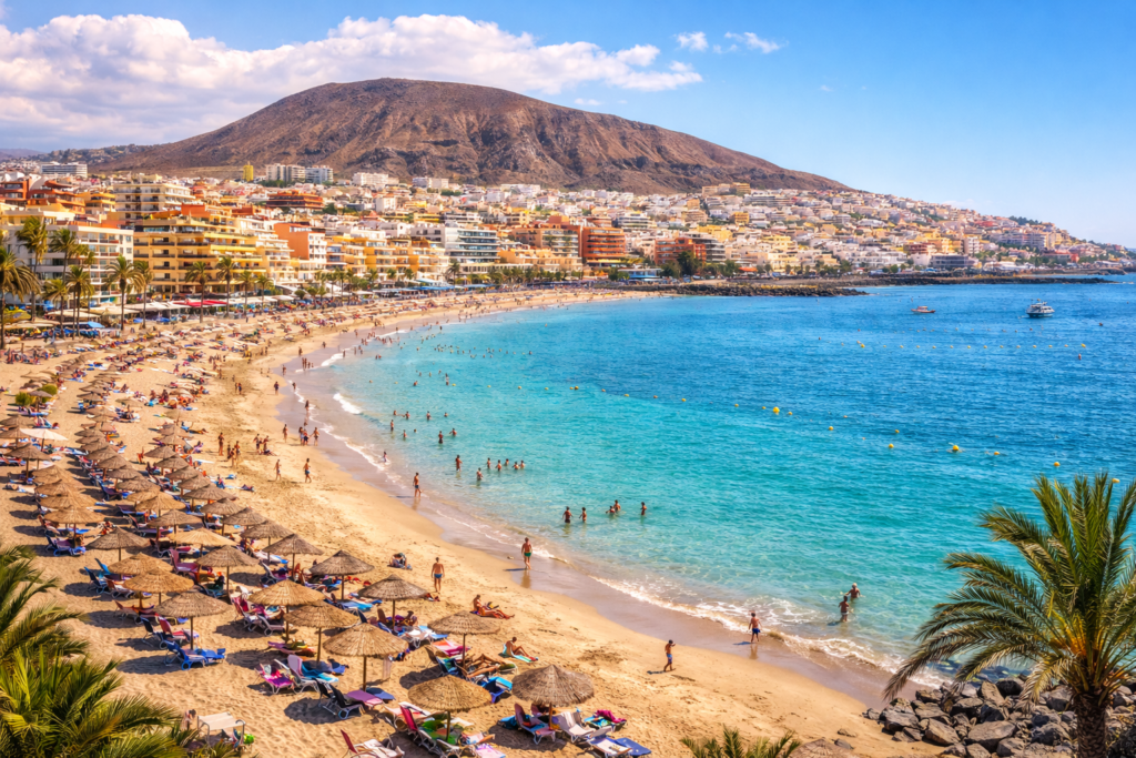 Tenerife Resort Guide 2026: Best Areas, Beaches & Things to Do 3 Beach at Los Cristianos in Tenerife with golden sand, calm turquoise water, sun loungers and parasols, a busy promenade, and hillside buildings in the background.